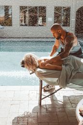 Tattooed male masseur giving an outdoor poolside back massage to a woman on a portable table beside a sunlit resort-style pool, spa relaxation scene.