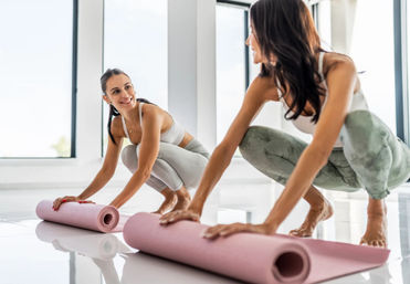 Two women rolling out pink yoga mats in a bright, sunlit studio, smiling as they prepare for a yoga/stretch session.