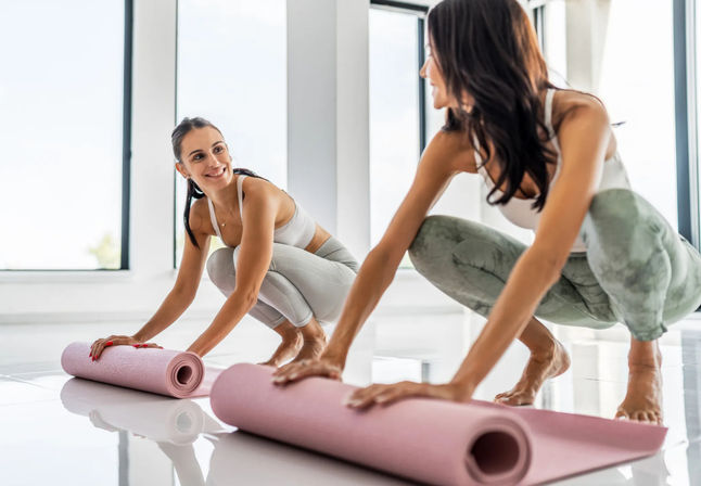 Two women rolling out pink yoga mats in a bright, sunlit studio, smiling as they prepare for a yoga/stretch session.