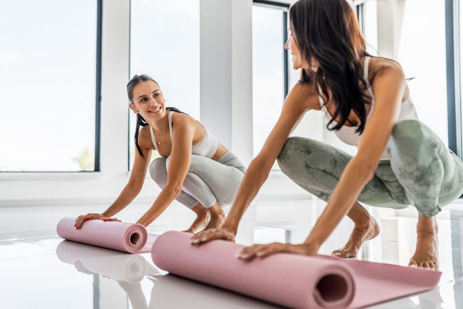 Two women in activewear smiling as they roll out pink yoga mats on the shiny floor of a bright sunlit fitness studio, preparing for yoga