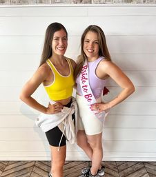 Two smiling women in athleisure posing indoors against a white shiplap wall — one in a yellow sports top and black bike shorts, the other in a lavender top, white shorts and a 'Bride to Be' sash for a casual bachelorette celebration.