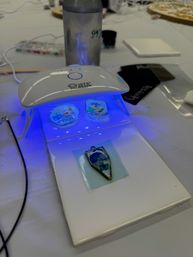 UV LED lamp curing blue resin floral pendants on a white craft table — DIY jewelry making station with tools and water bottle
