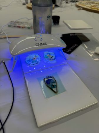 UV LED lamp curing blue resin floral pendants on a white craft table — DIY jewelry making station with tools and water bottle