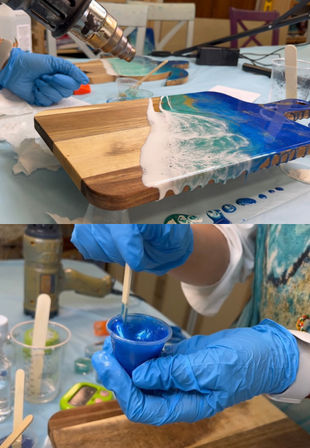 Close-up of DIY resin art: gloved hands stirring bright blue epoxy and using a heat gun to create ocean wave effects on a wooden cutting board.