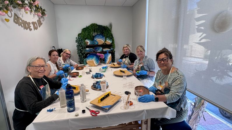 Smiling group of six women in aprons and blue gloves around a long table in a bright craft studio, making ocean‑themed resin wood boards and trays with cups, tools and paint scattered — lively bachelorette craft party scene.