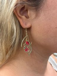 Close-up of an ear wearing a gold leaf-shaped dangle earring with two bright pink floral accents, blonde hair framing the lobe — boho jewelry close-up.