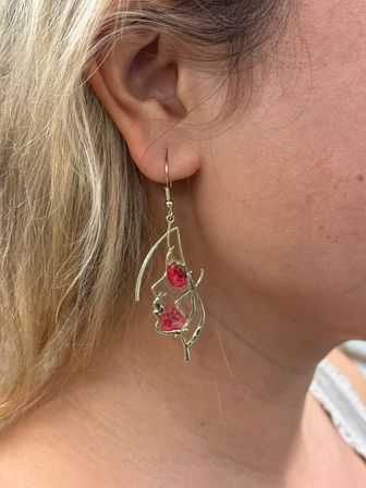 Close-up of an ear wearing a gold leaf-shaped dangle earring with two bright pink floral accents, blonde hair framing the lobe — boho jewelry close-up.
