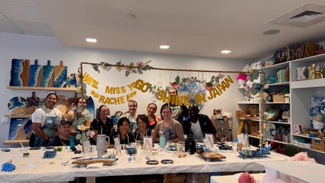 Smiling group photo in an indoor art studio at a resin workshop, ocean-themed boards on shelves, long table with paints, tools and a farewell banner overhead.