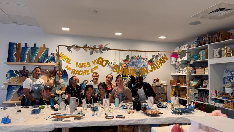 Smiling group photo in an indoor art studio at a resin workshop, ocean-themed boards on shelves, long table with paints, tools and a farewell banner overhead.