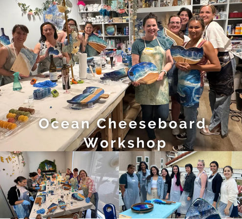 Smiling group of participants in an indoor art studio holding handmade ocean-themed resin-and-wood cheeseboards at an Ocean Cheeseboard Workshop, surrounded by paint supplies, drinks and snacks.