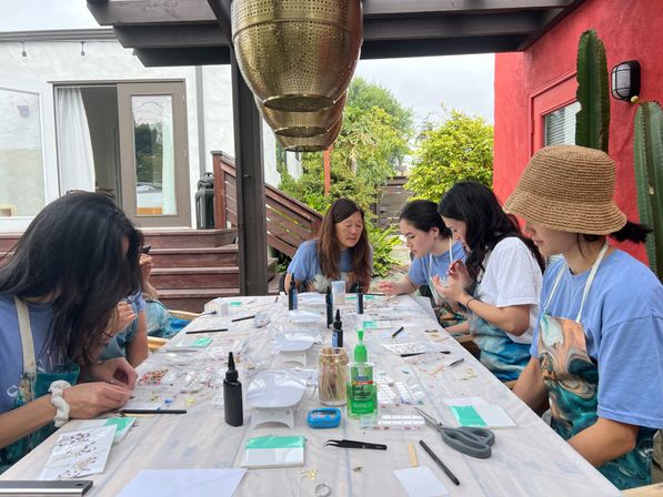 Group of people at an outdoor patio craft workshop, seated around a long table under a wooden pergola with beads, tweezers, small trays, glue bottles and hand sanitizer, red stucco wall and cactus in the background.