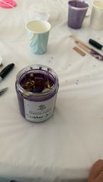 Purple scented candle in a glass jar topped with dried flowers and a wooden wick on a white tablecloth surrounded by paper cups and crafting tools — DIY candle-making setup.