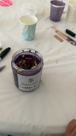 Purple scented candle in a glass jar topped with dried flowers and a wooden wick on a white tablecloth surrounded by paper cups and crafting tools — DIY candle-making setup.