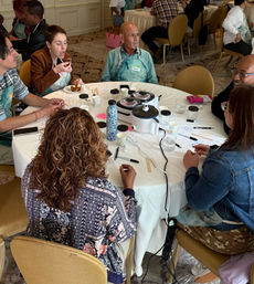 Hands-on group workshop in a hotel conference room: six people in aprons around a round table with small wax warmers, cups, wooden spatulas and snacks.