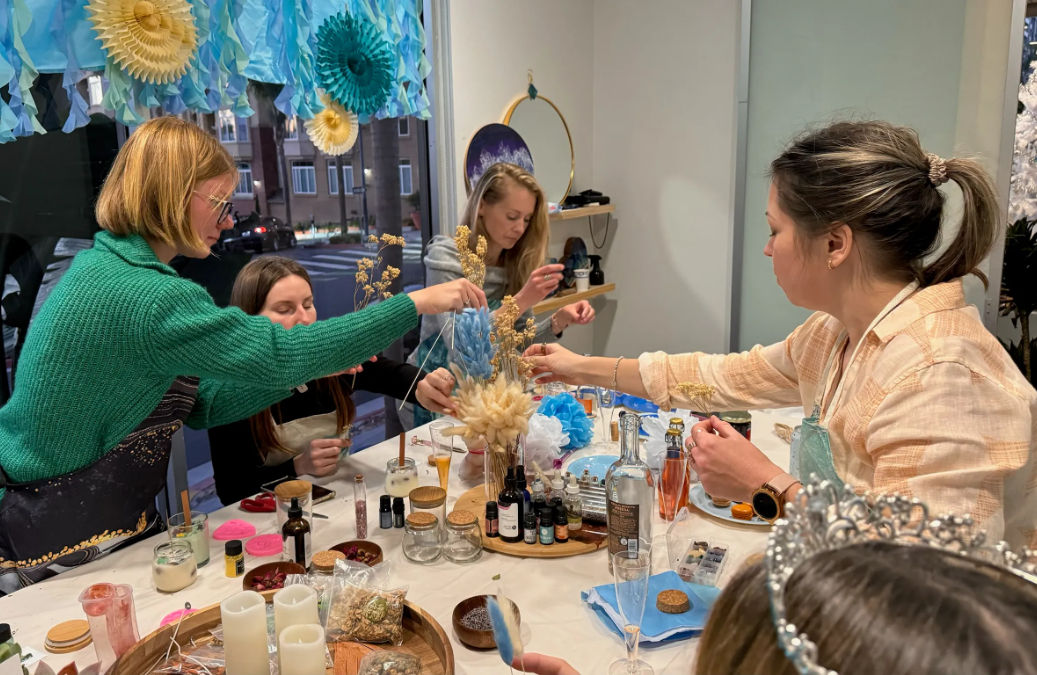 Group of women at an indoor DIY craft workshop arranging dried flowers and supplies on a cluttered table beneath blue paper decorations by a street-facing window.