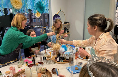 Group of women at an indoor DIY craft workshop arranging dried flowers and supplies on a cluttered table beneath blue paper decorations by a street-facing window.