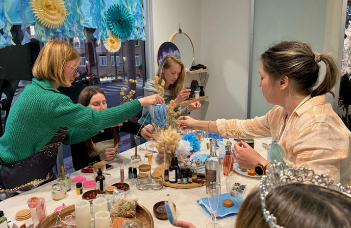 Group of women at an indoor DIY craft workshop arranging dried flowers and supplies on a cluttered table beneath blue paper decorations by a street-facing window.