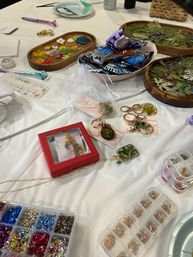Colorful indoor craft workshop table with DIY resin jewelry supplies: wooden trays of pressed flowers and leaves, assorted keychain and pendant charms, small containers of glitter and metal jump rings, and a bowl of snacks on a white tablecloth.