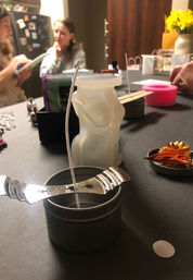 DIY candle-making table with a figure-shaped wax mold and a wick centered in a metal tin using a wick holder, dried flower petals and craft tools with blurred people in the background.