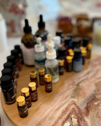 Assortment of essential oil vials and glass dropper bottles with gold caps arranged on a round wooden tray over a pink marble countertop — spa-style aromatherapy setup.