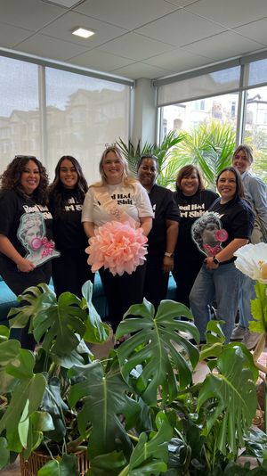 Smiling group of seven women inside a sunlit indoor plant shop celebrating a bridal shower; bride-to-be wears a sash and tiara and holds a large pink paper pom-pom, with lush monstera and potted plants in the foreground.