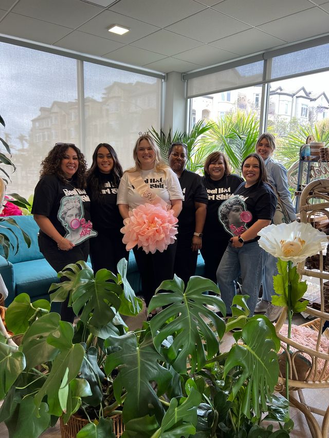 Seven women pose in a bright modern lounge for a bachelorette photo — bride-to-be in sash and tiara holding a large pink pom-pom, friends in matching graphic tees, surrounded by tropical houseplants and large windows.