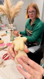 Close-up of hands holding a cream-colored, torso-shaped candle with a wick during a DIY candle-making workshop, blurred participant, tools and pampas grass vase in the background.