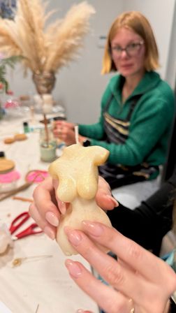 Close-up of hands holding a cream-colored, torso-shaped candle with a wick during a DIY candle-making workshop, blurred participant, tools and pampas grass vase in the background.