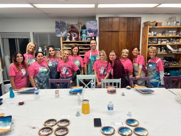 Smiling group of adults in matching pink shirts at an indoor craft studio paint party, standing behind a table with blue-and-white resin coasters, trays, paint supplies and water bottles.