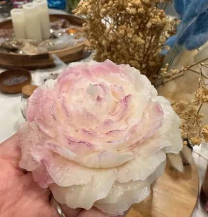 Hand holding a pink-and-white rose-shaped wax candle with layered petals and a subtle glittery sheen, dried flowers and a wooden tray blurred in the background — decorative tabletop decor.