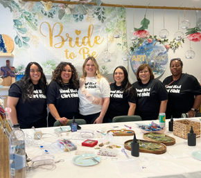 Smiling group of six women at a bachelorette craft workshop in an indoor studio, bride-to-be wearing a sash in front of a gold “Bride to Be” banner, table covered with DIY jewelry-making supplies and floral decorations.