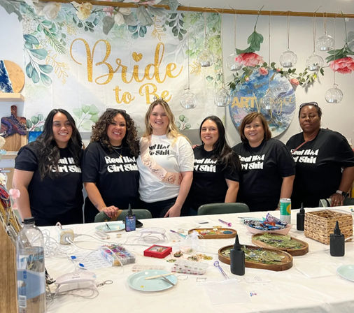 Smiling group of six women at a bachelorette craft workshop in an indoor studio, bride-to-be wearing a sash in front of a gold “Bride to Be” banner, table covered with DIY jewelry-making supplies and floral decorations.
