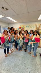 Group of eight women in a bright art studio wearing aprons and floral leis, smiling and holding handmade jewelry and resin pieces after a fun craft workshop.