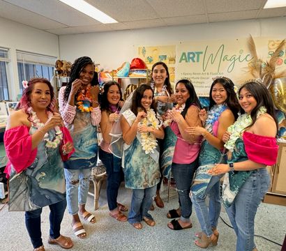 Group of eight women in a bright art studio wearing aprons and floral leis, smiling and holding handmade jewelry and resin pieces after a fun craft workshop.