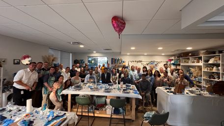 Energetic corporate team-building art workshop in an indoor studio — about 40 people in aprons posing around long tables with blue resin supplies and a 'Happy Valentine's Day' heart balloon overhead.