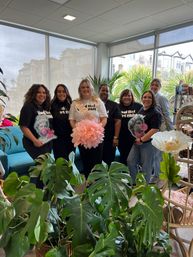 Group of seven women smiling at a bachelorette celebration inside a bright, plant-filled boutique with large windows, tropical greenery, oversized paper flowers and cozy seating.
