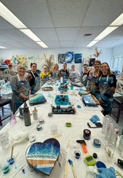 Group of people in a bright art studio gathered around a long table during a resin ocean art workshop, displaying blue-and-white coastal-inspired resin wave paintings on wood (including a heart-shaped panel) with cups, stir sticks, torches and craft supplies scattered across the table.