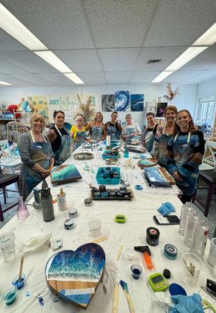 Group of people in a bright art studio gathered around a long table during a resin ocean art workshop, displaying blue-and-white coastal-inspired resin wave paintings on wood (including a heart-shaped panel) with cups, stir sticks, torches and craft supplies scattered across the table.
