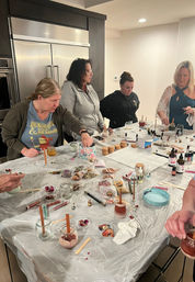 Group of women around a kitchen island making DIY candles — jars with wooden wicks, dried flowers, bottles of oils and craft supplies spread across a plastic-covered table.