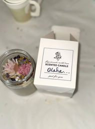 Top-down view of a handmade scented candle in a glass jar with pink flower-shaped wax decorations and crystals, beside a white gift box labeled "Scented Candle Aloha" on a white table.