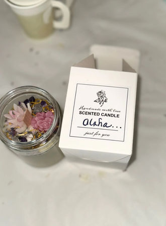 Top-down view of a handmade scented candle in a glass jar with pink flower-shaped wax decorations and crystals, beside a white gift box labeled "Scented Candle Aloha" on a white table.