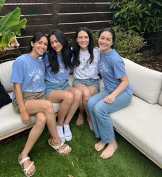 Four smiling friends wearing San Diego T-shirts and casual summer outfits seated on a white outdoor patio couch in a backyard with a wooden fence and green plants.