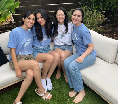 Four smiling friends wearing San Diego T-shirts and casual summer outfits seated on a white outdoor patio couch in a backyard with a wooden fence and green plants.