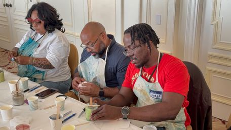 Three adults in aprons concentrating at a bright indoor art workshop, mixing paint in cups and using wooden sticks, brushes and tools on a table.