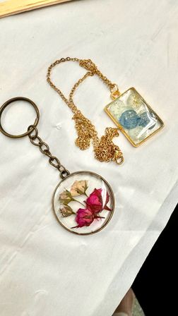 Botanical resin jewelry: round keychain with preserved pink and white rosebuds and bronze ring, paired with a gold-chain rectangular pendant featuring a blue skeleton leaf and gold flakes on white fabric.
