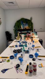 Indoor craft studio table set for a birthday resin art workshop — white-covered workspace with resin bottles, pigments, gloves, mixing cups, tape, levels and tools, champagne flutes, and blue ocean-style resin boards on a green foliage backdrop.