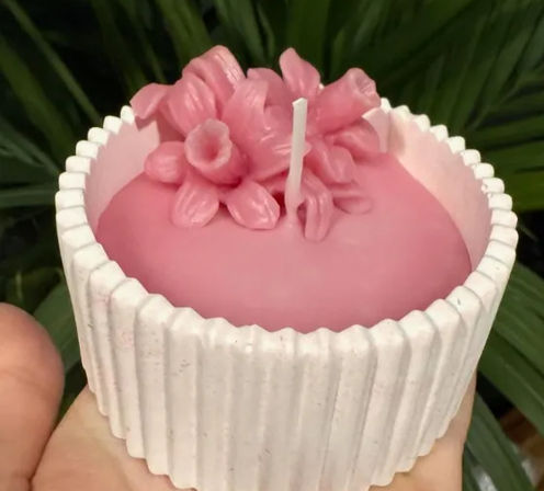 Handheld decorative pink wax candle topped with sculpted floral petals and a single wick, set in a white ribbed ceramic holder against a leafy green background
