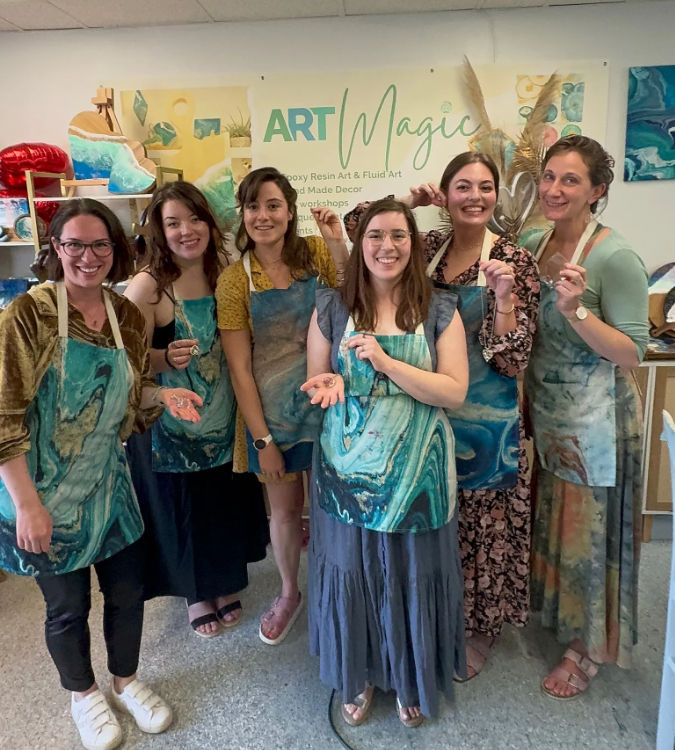 Six women smiling in an art studio, wearing teal marbled aprons and holding small epoxy resin pieces after a hands-on fluid-art workshop, colorful resin artwork and supplies visible in the background.