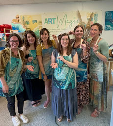 Six women smiling in an art studio, wearing teal marbled aprons and holding small epoxy resin pieces after a hands-on fluid-art workshop, colorful resin artwork and supplies visible in the background.