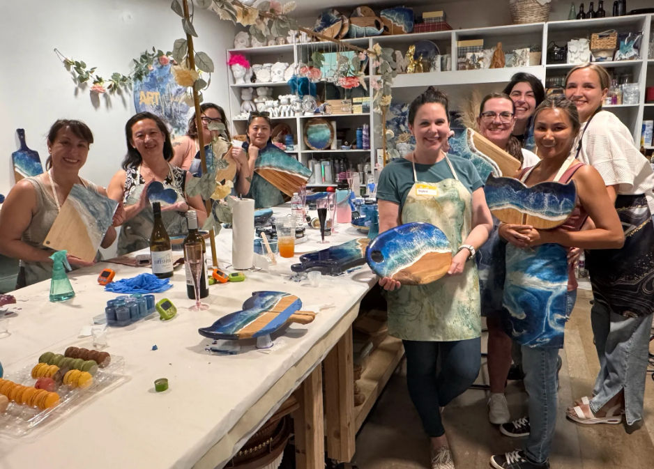 Smiling group of women in an art studio holding blue-green resin “ocean” wooden serving boards at a DIY resin art workshop, paint-covered table with tools, drinks and snacks.